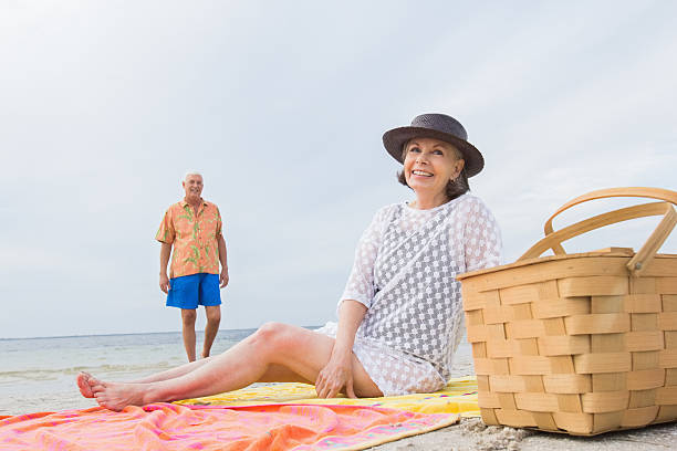 Woman over 60 in stylish beachwear sitting on a blanket, with a man and picnic basket nearby.